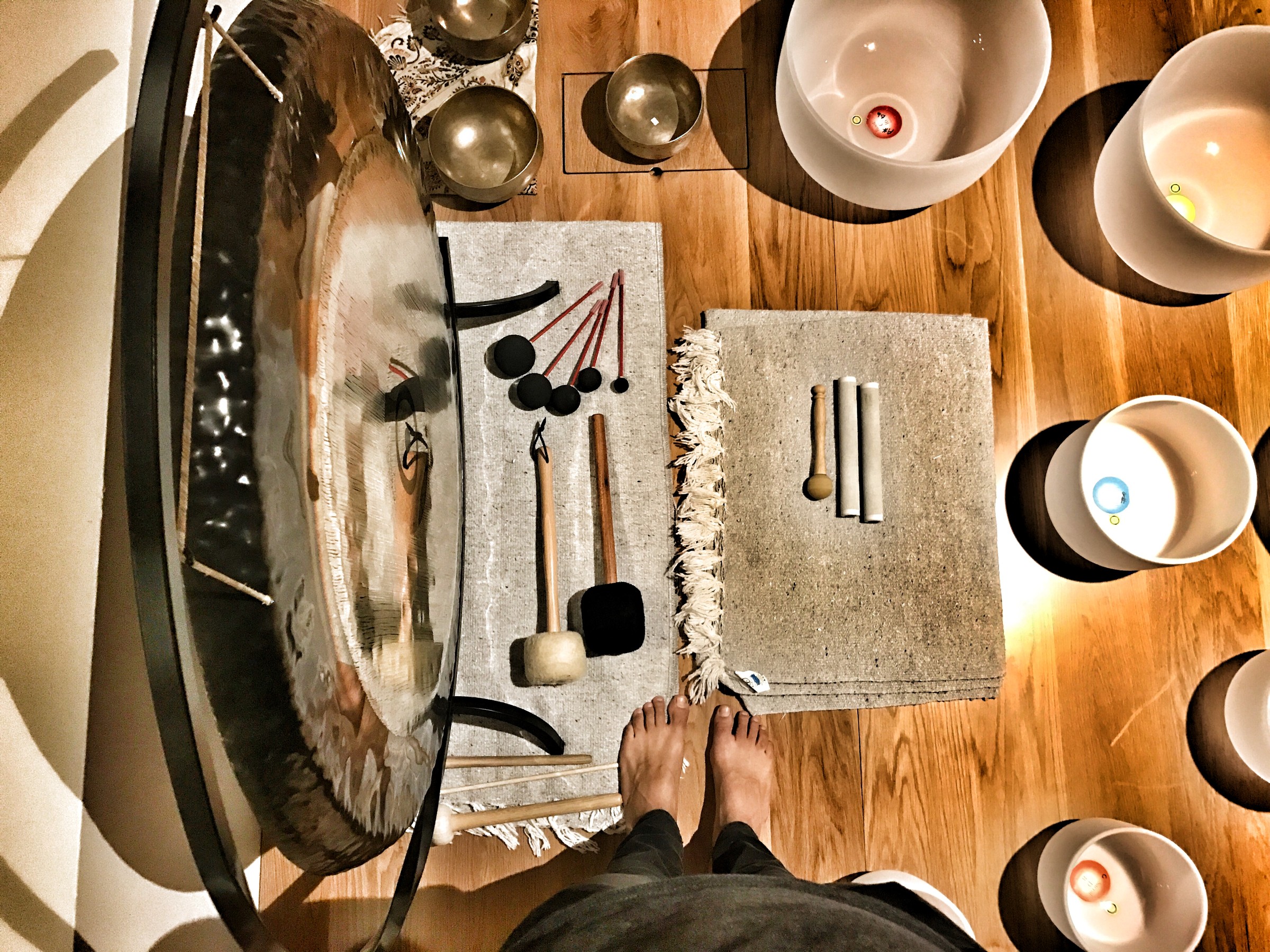 Looking down on a gong on its stand, mallets set out on a mat beside it, metal bowls, crystal bowls holding lit candles, and a player's bare feet