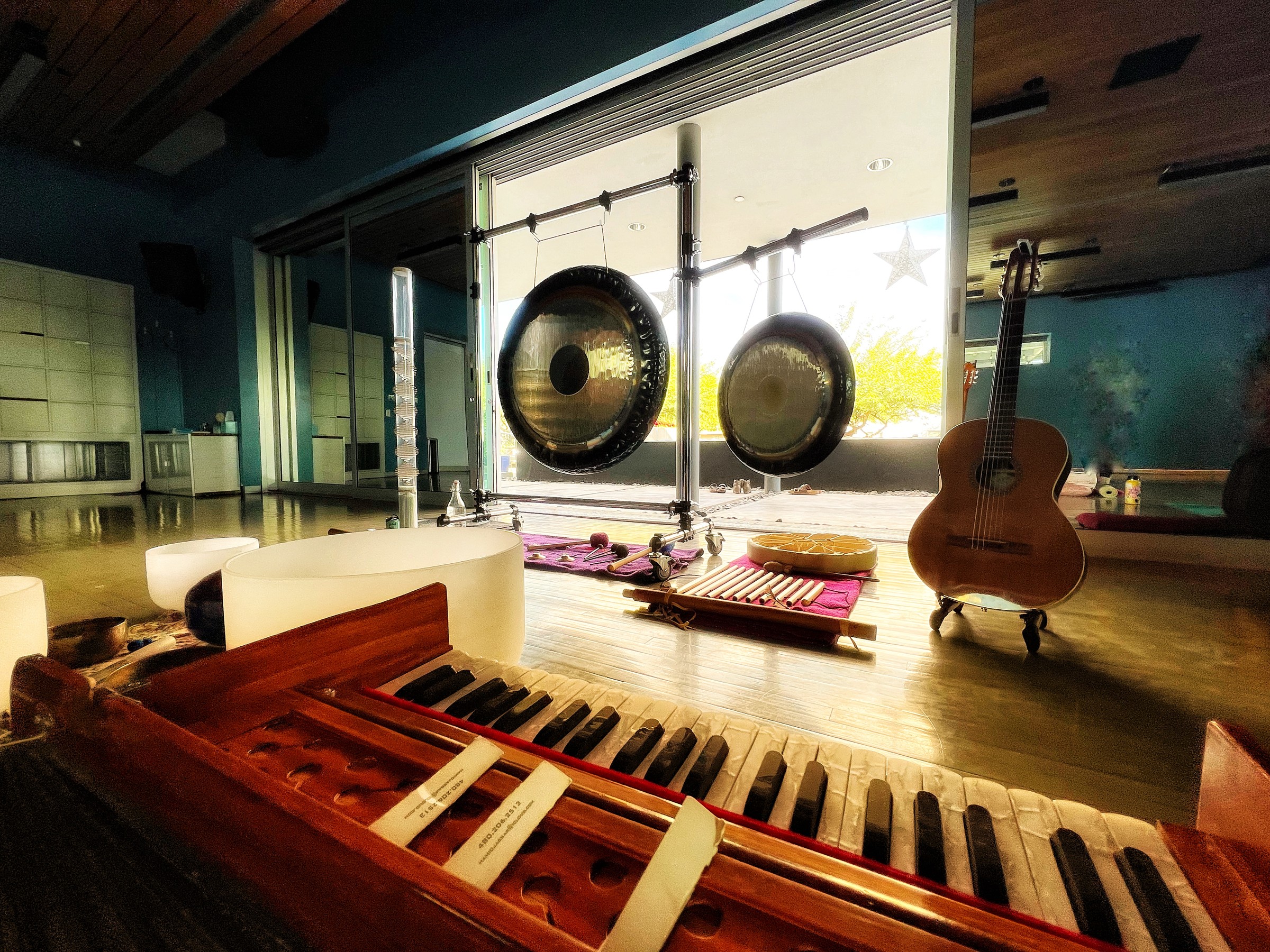 Two gongs on stands by a window, with a guitar, crystal singing bowls and a harmonium