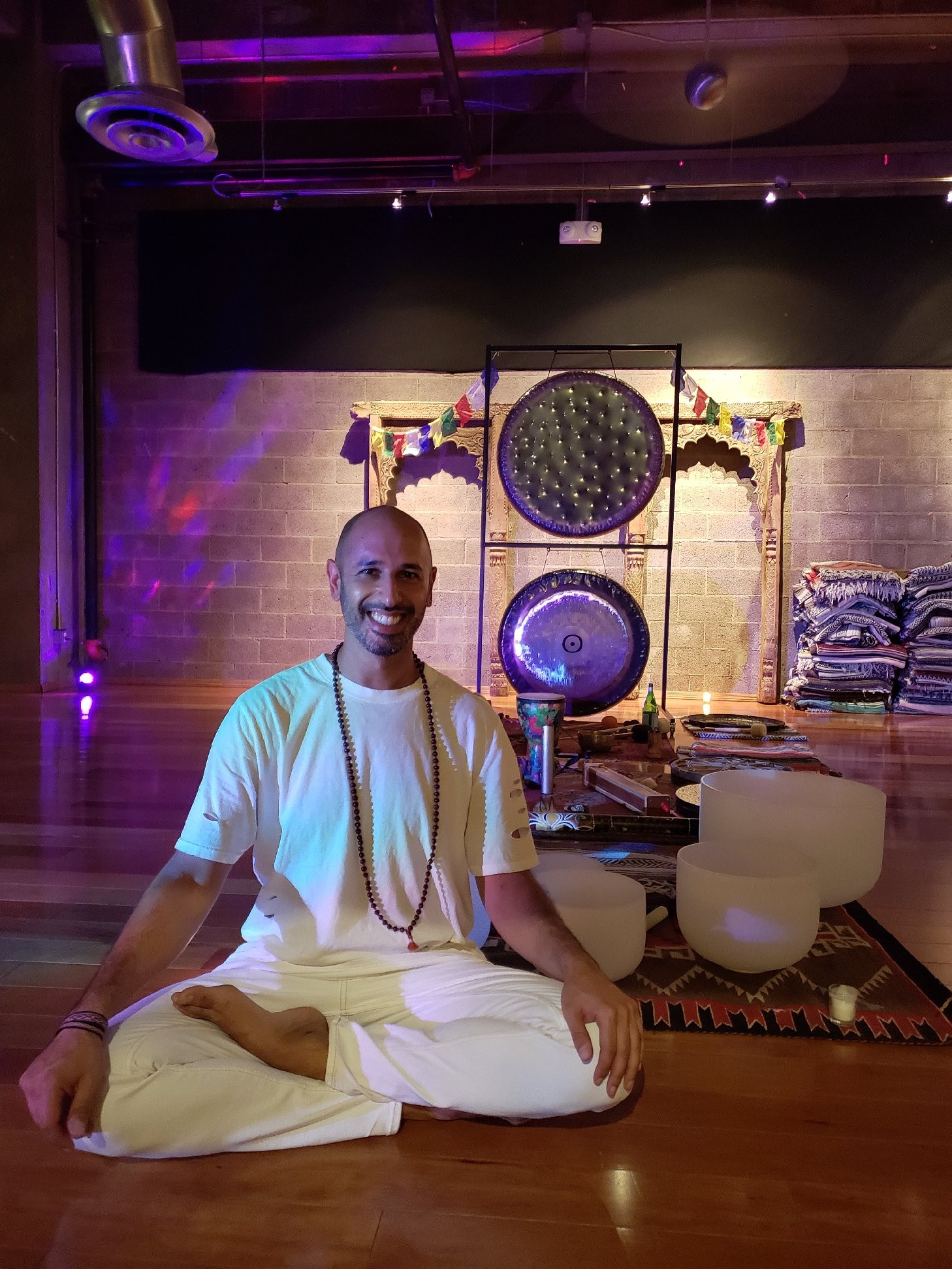 Hamid Jabbar sitting cross-legged in white, with two gongs on a stand and a row of crystal bowls behind him
